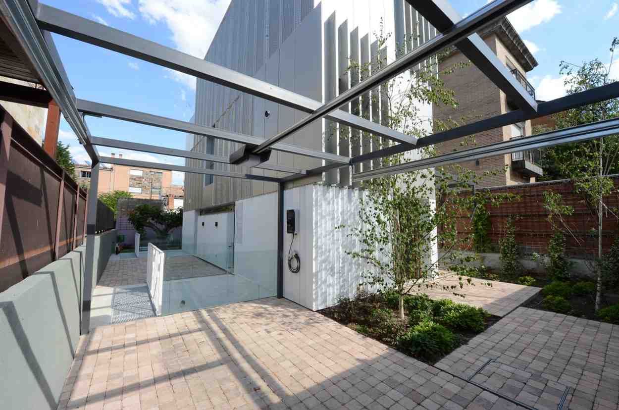 Patio moderno con paredes blancas, vigas de pérgola, adoquines y parterres junto a un edificio.