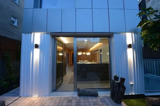 Entrada moderna a la tienda con fachada blanca, puertas de cristal e iluminación interior cálida al anochecer.