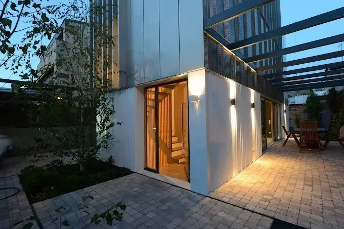 Exterior de una casa moderna al anochecer con entrada iluminada, paredes blancas, pavimento de patio.