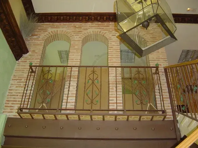 Balcón interior con barandillas ornamentadas, ventanas arqueadas y una lámpara de techo colgante de cristal.