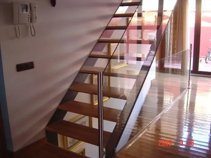 Escalera interior moderna con escalones de madera y barandilla de cristal junto a una habitación soleada.