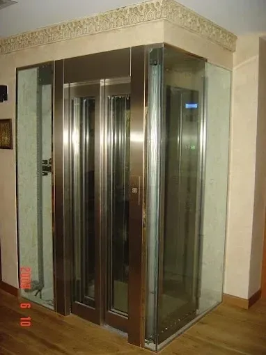 Ascensor de cristal en un rincón de una casa con puertas de espejo y suelo de madera.