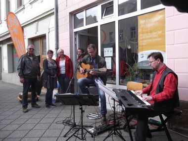 Ein Musikduo tritt auf dem Bürgersteig vor einem Laden auf, während drei Personen stehen und zusehen.