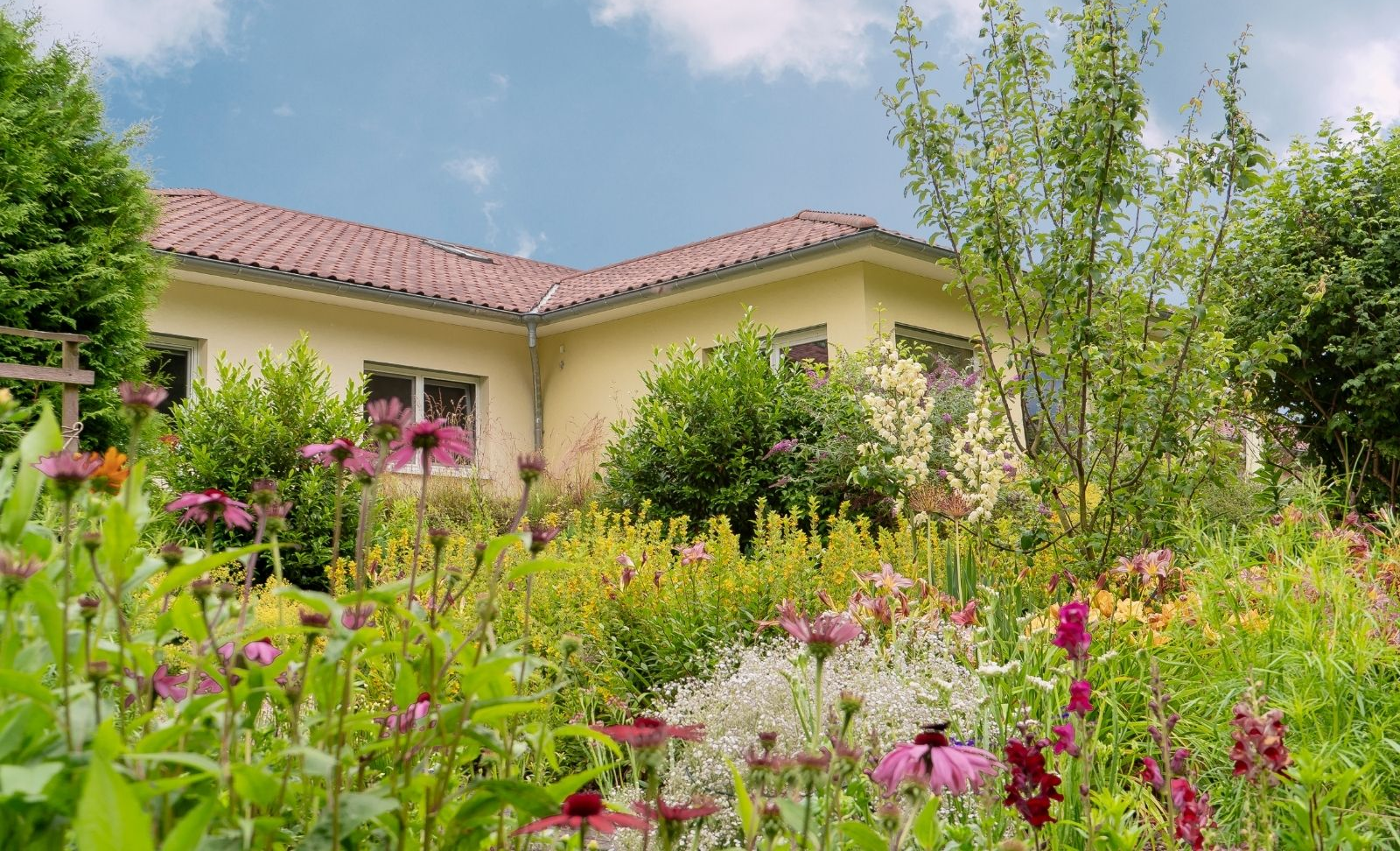 Ein gelbes Haus mit rötlichem Dach hinter einem üppigen, farbenfrohen Blumengarten unter blauem Himmel.