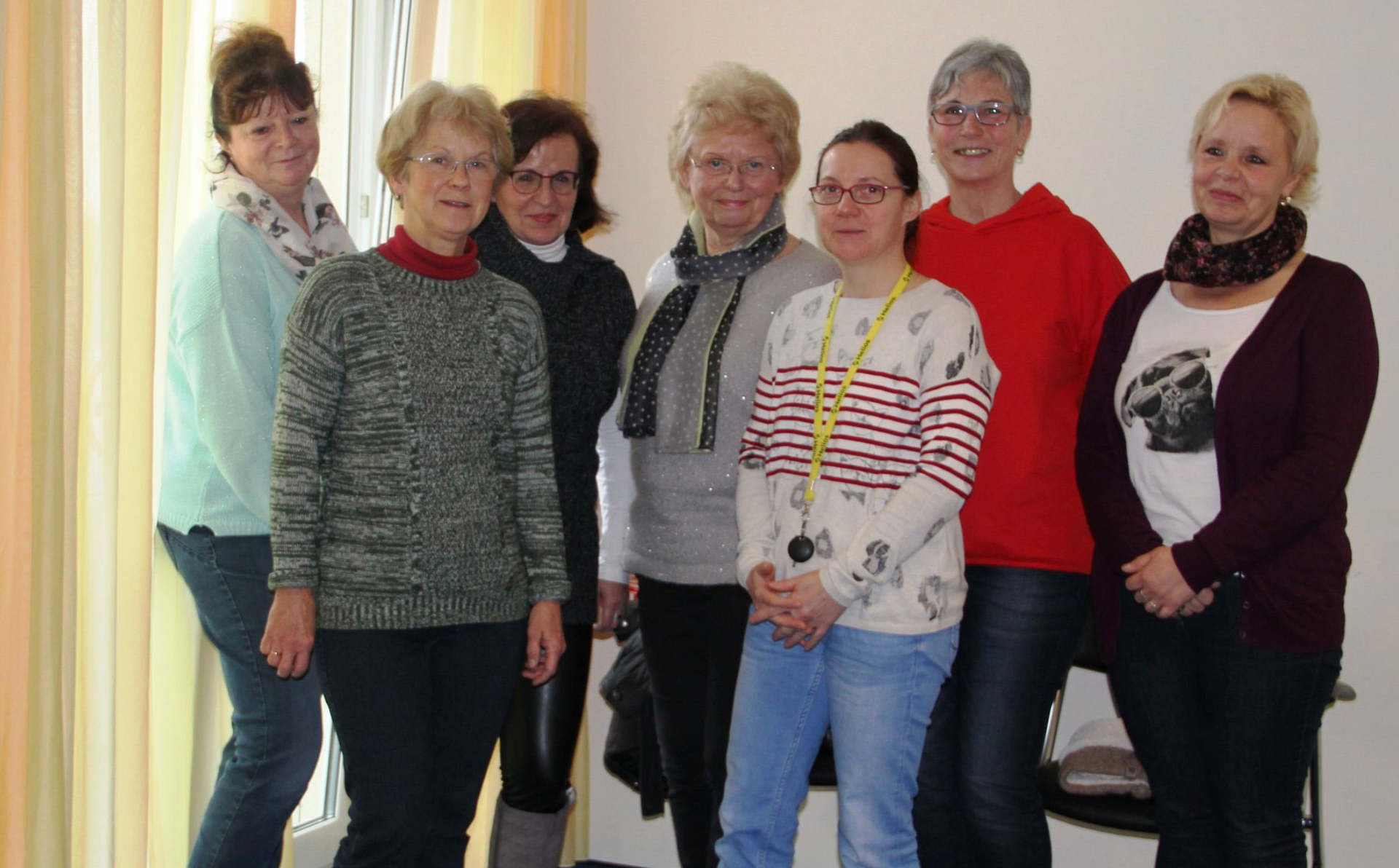 Sieben Hospizbegleiter posieren für ein Gruppenfoto vor einer schlichten Wand, gekleidet in lässige Pullover und Oberteile.
