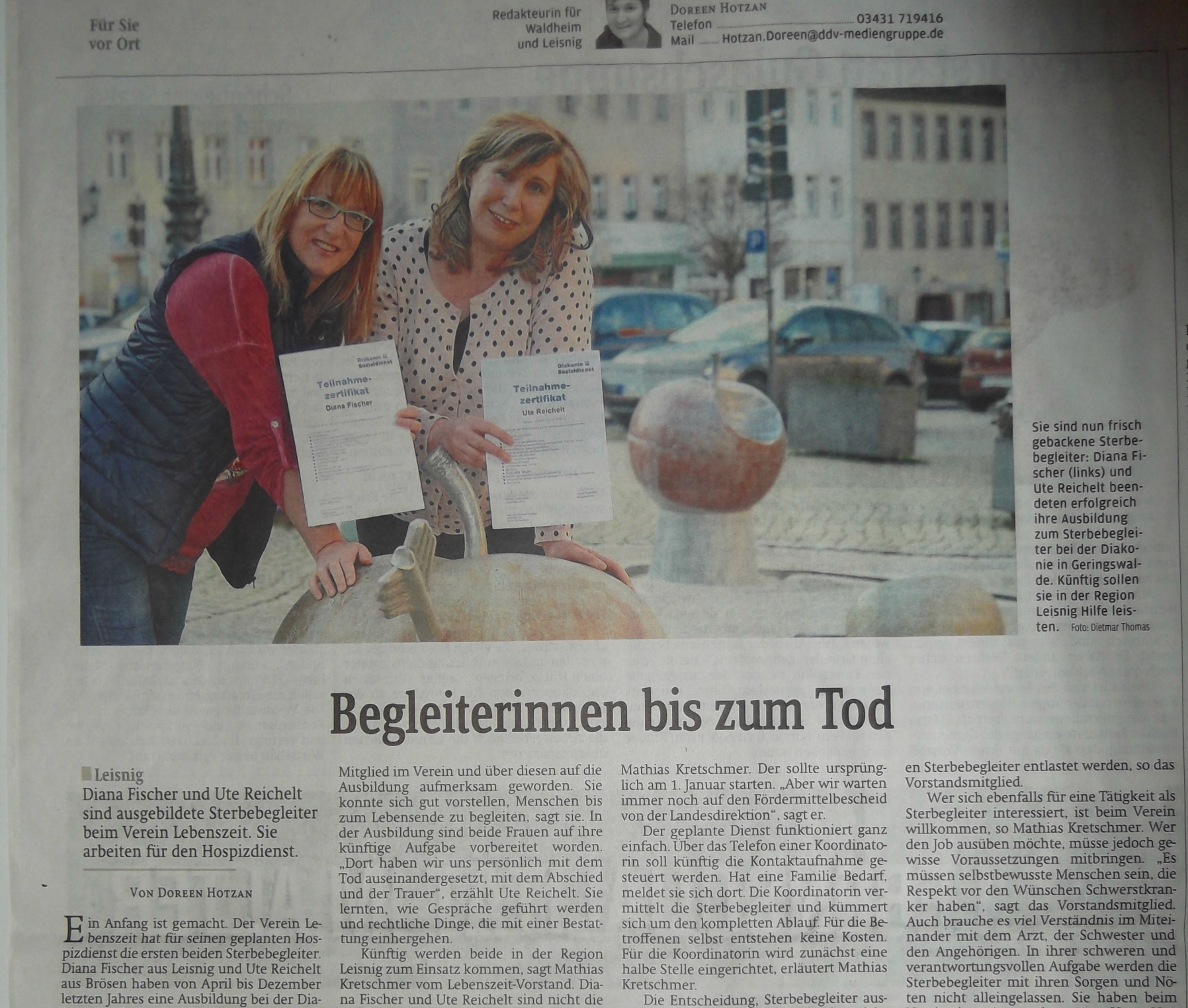 Zwei lächelnde Menschen stehen auf einem Stadtplatz und halten Zertifikate zur Sterbebegleitung in den Händen.