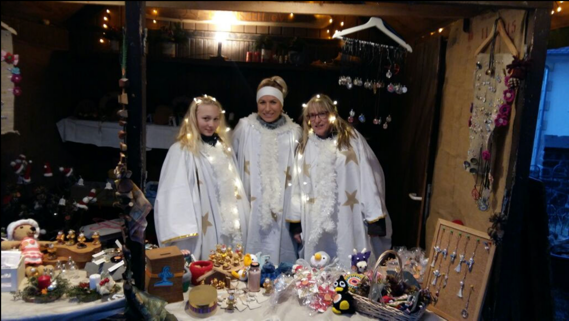 Drei Personen in weißen, mit Lichtern und goldenen Sternen verzierten Umhängen stehen hinter einem Marktstand.