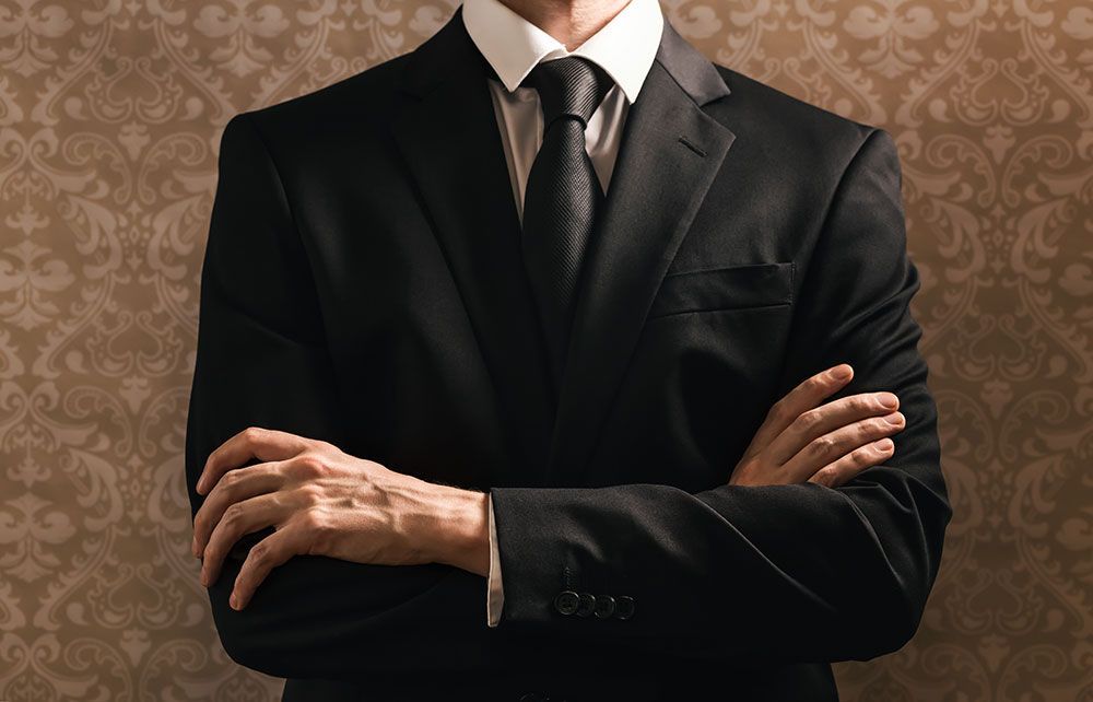 Mann in schwarzem Anzug und Krawatte mit verschränkten Armen vor gemustertem Hintergrund.