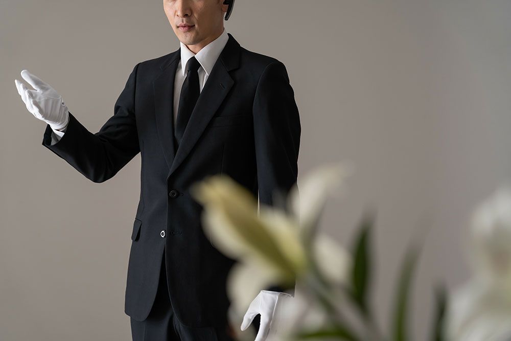 Mann im schwarzen Anzug, mit weißen Handschuhen, gestikuliert in Richtung weißer Blumen.