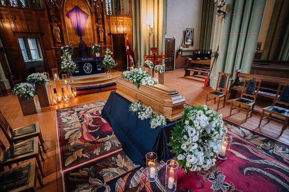 Trauerfeier in einer Kirche mit einem marineblau gehüllten Holzsarg, umgeben von weißen Blumen und Kerzen.
