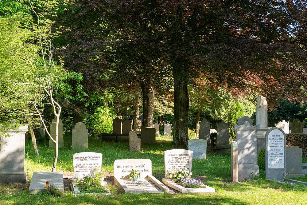 Friedhof mit Grabsteinen unter Bäumen, sonniger Tag.