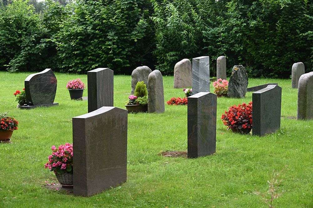 Friedhofsszene mit Grabsteinen auf grünem Gras; bunte Blumen in Töpfen und Beeten; Bäume im Hintergrund.