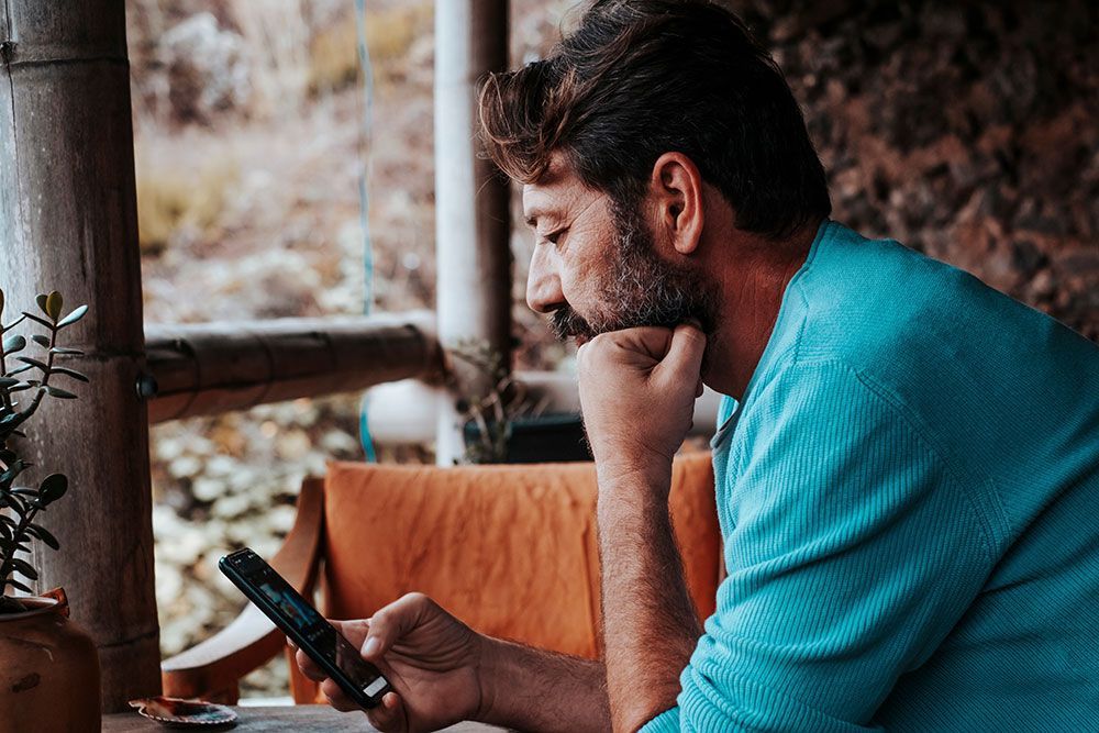 Mann mit Bart schaut auf den Bildschirm seines Telefons, draußen auf einer Veranda, und stützt sein Kinn auf seine Hand.