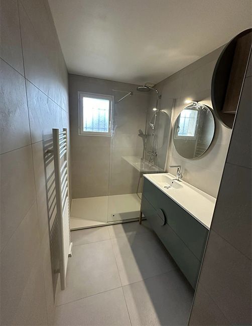 Salle de bains moderne avec douche à l’italienne, carrelage gris, miroir mural et meuble-lavabo blanc.