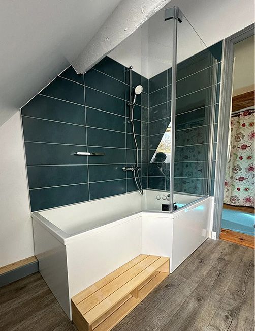 Salle de bains moderne avec baignoire blanche, douche carrelée foncée, paroi vitrée et banc en bois sous un plafond mansardé
