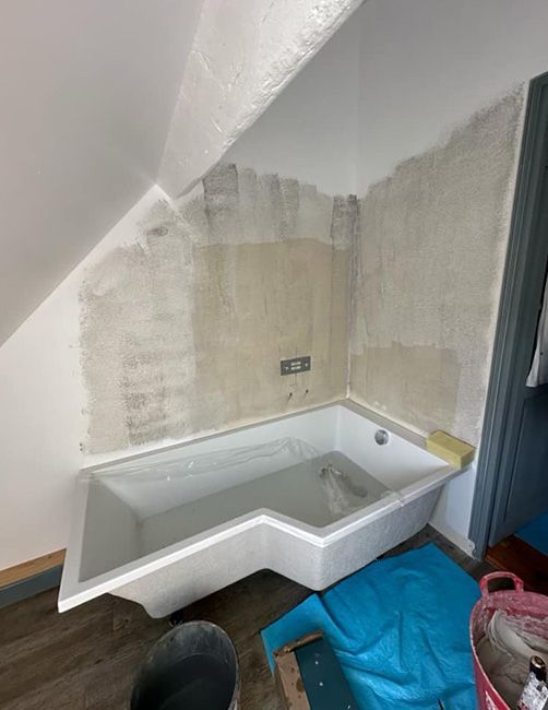 Petite salle de bains d’angle avec une baignoire blanche sous un plafond mansardé et des murs bruts.