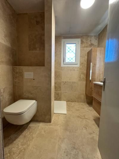 Salle de bains moderne beige avec toilettes suspendues, douche à l’italienne, fenêtre et porte vitrée à droite