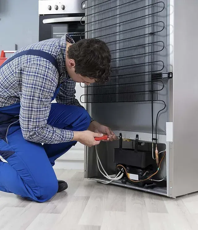 Un hombre con un mono azul repara el compresor del refrigerador con un destornillador.