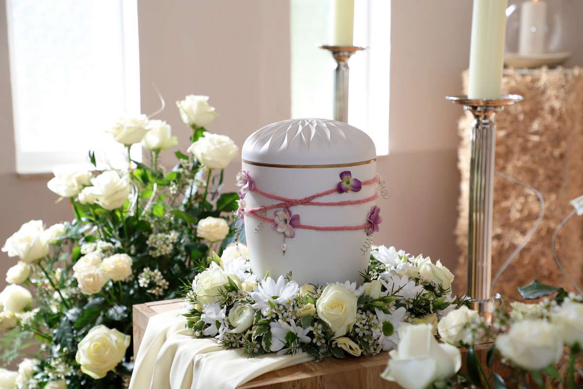 In einer Kirche ist eine weiße Urne von weißen Blumen und Kerzen umgeben.