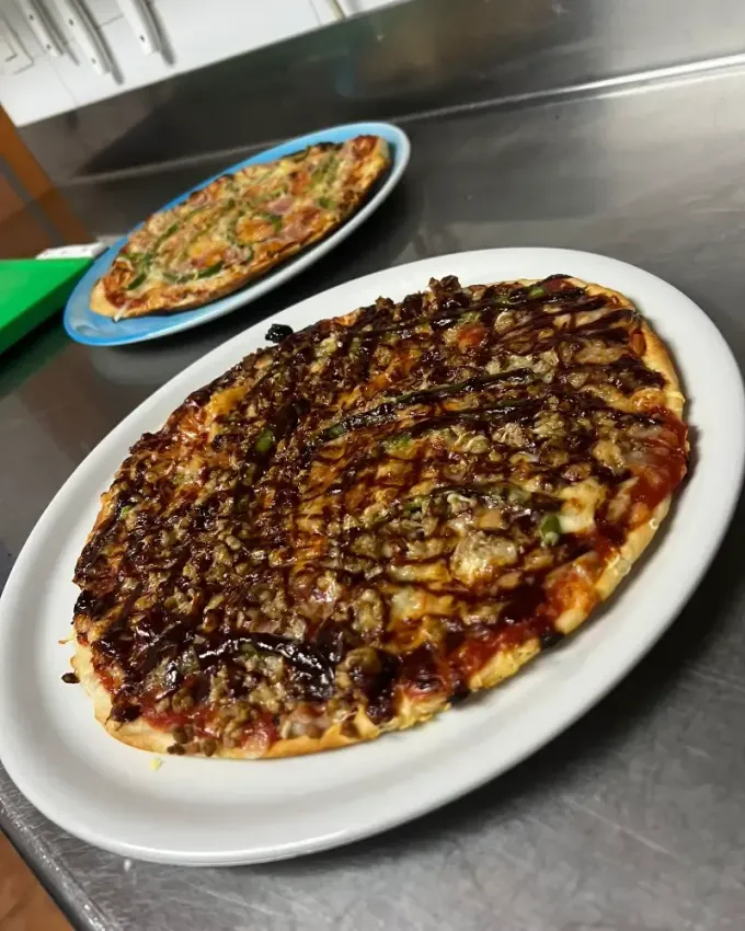 Dos pizzas en platos blancos sobre un mostrador de acero inoxidable, una de ellas cubierta con glaseado oscuro.