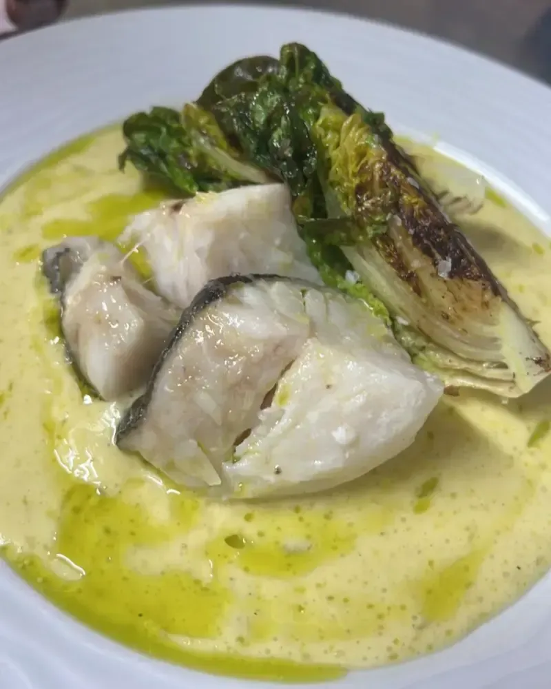 Pescado emplatado con verduras asadas en salsa amarilla sobre un plato blanco.