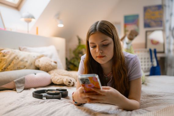 Un adolescent allongé sur son lit utilise un smartphone. On aperçoit un ours en peluche et un collier pour animal de compagnie.
