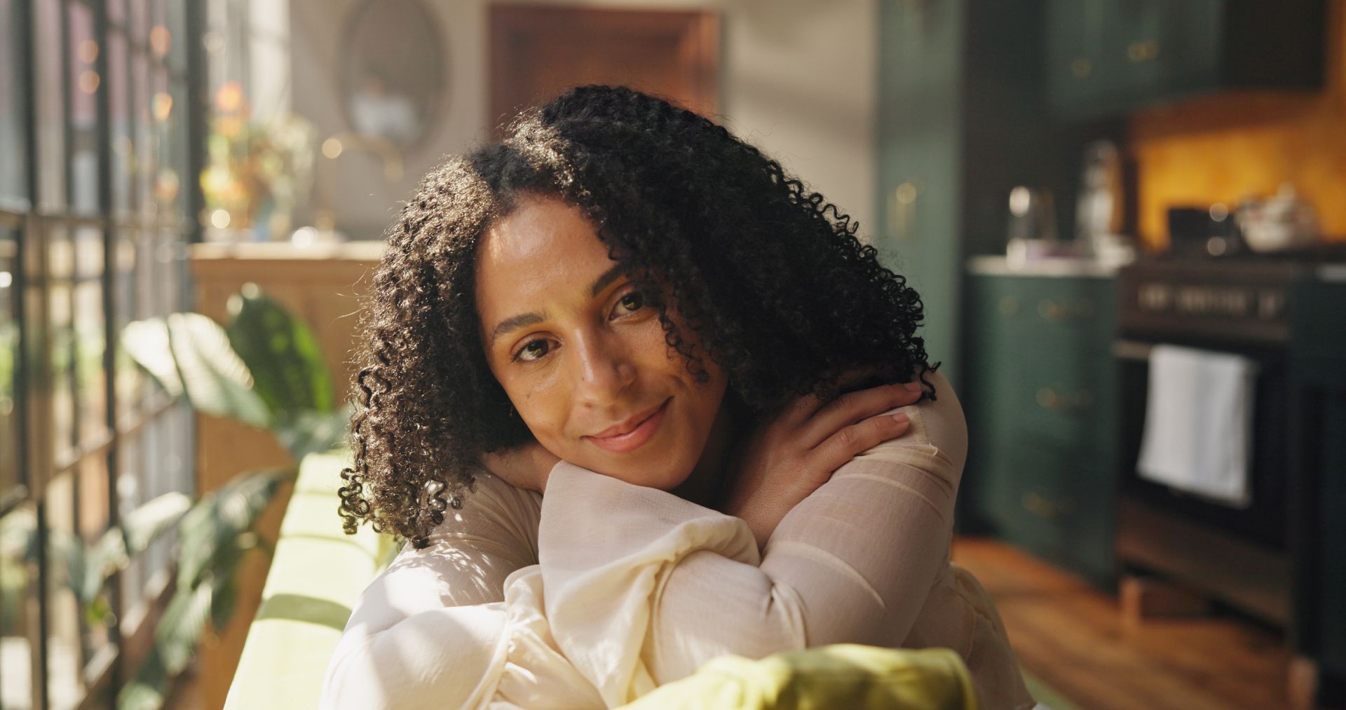 Une femme aux cheveux bouclés sourit, appuyée sur un rebord de fenêtre ensoleillé ; la cuisine est visible en arrière-plan.