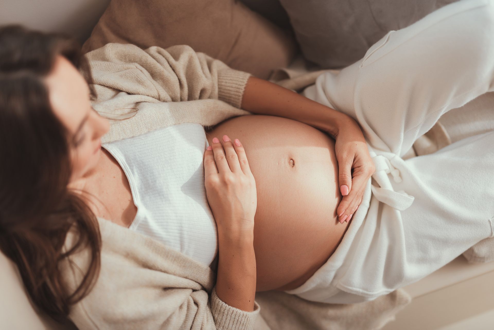 Femme enceinte se reposant, les mains sur le ventre, vêtue de blanc, à l'intérieur.