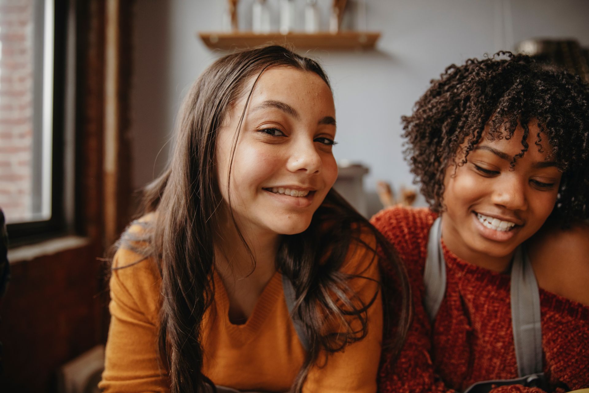 Deux jeunes gens sourient, l'un avec des tresses et un appareil dentaire, l'autre avec des cheveux bouclés, dans une pièce avec une fenêtre.