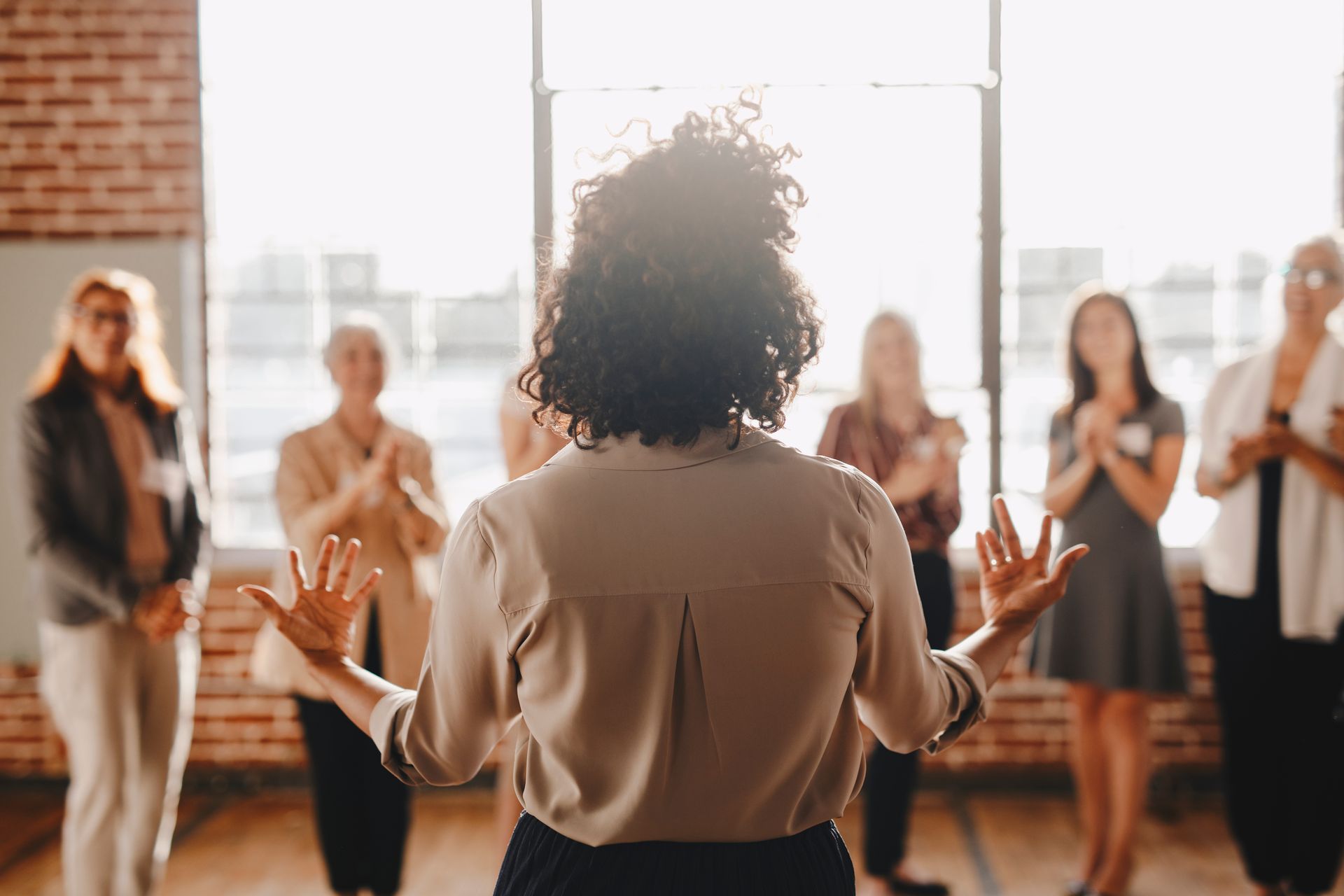 Femme s'adressant à un groupe qui applaudit. Pièce ensoleillée, vue de l'arrière.