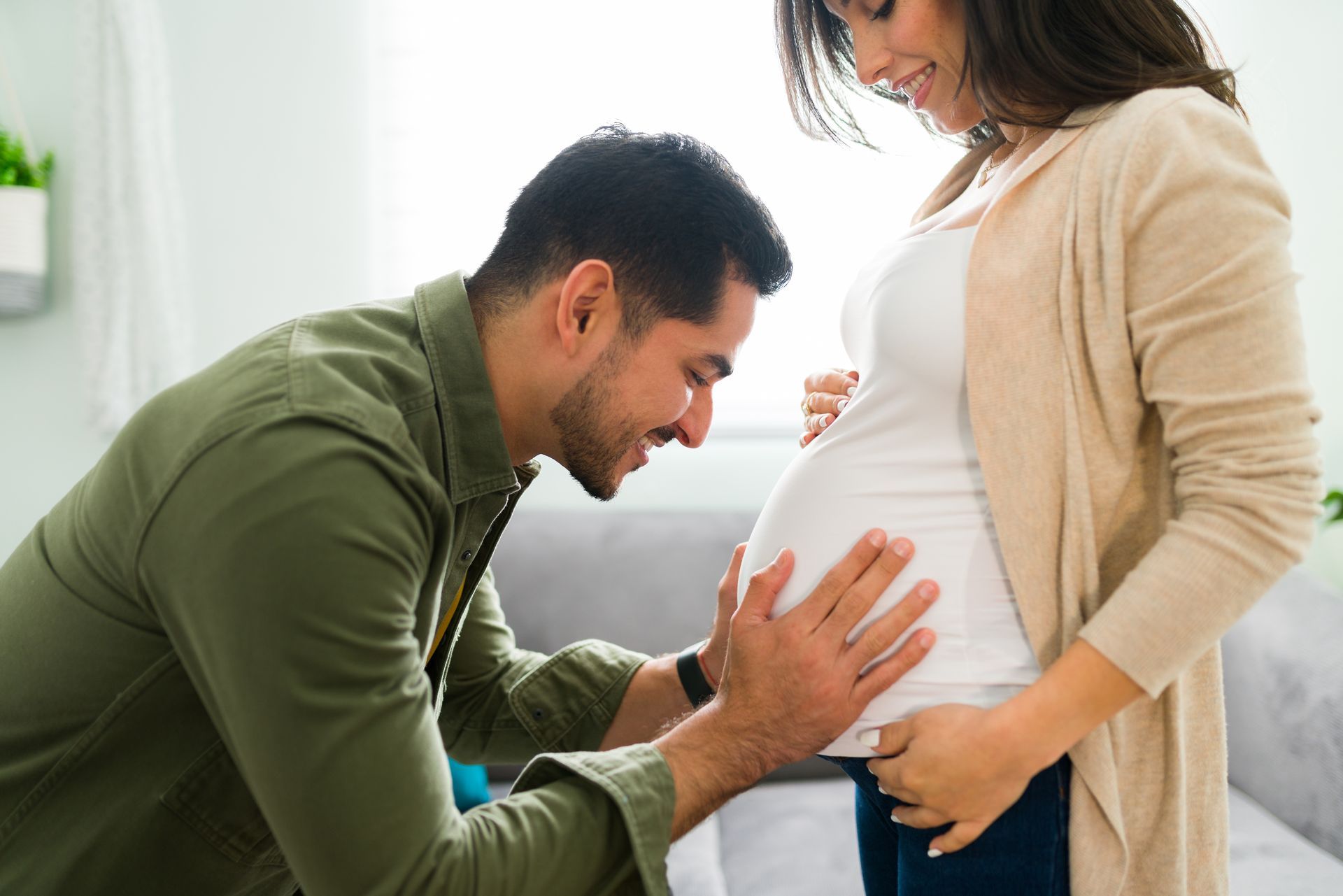 Un homme caresse le ventre d'une femme enceinte en souriant. La femme sourit, les mains sur son ventre. À l'intérieur, en plein soleil.