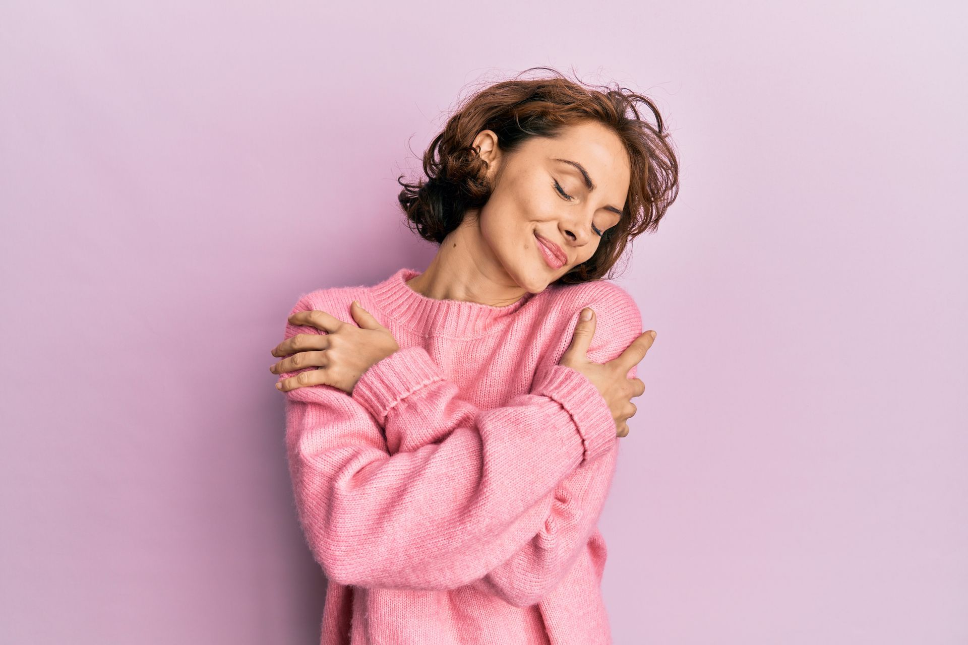 Une femme en pull rose se serre contre elle-même, les yeux fermés, souriante, sur un fond lavande.