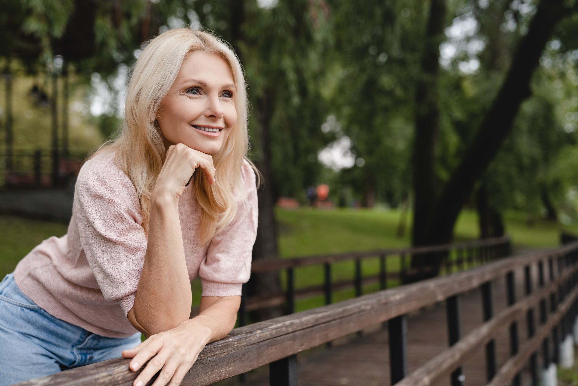 Une femme blonde, souriante, est appuyée sur une rambarde en bois. Elle porte un haut rose pâle et un jean. Le décor est celui d'un parc.