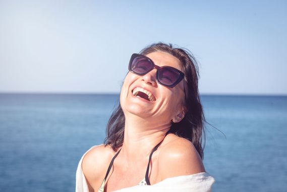 Femme riant en extérieur, portant des lunettes de soleil, avec l'océan et le ciel bleu en arrière-plan.