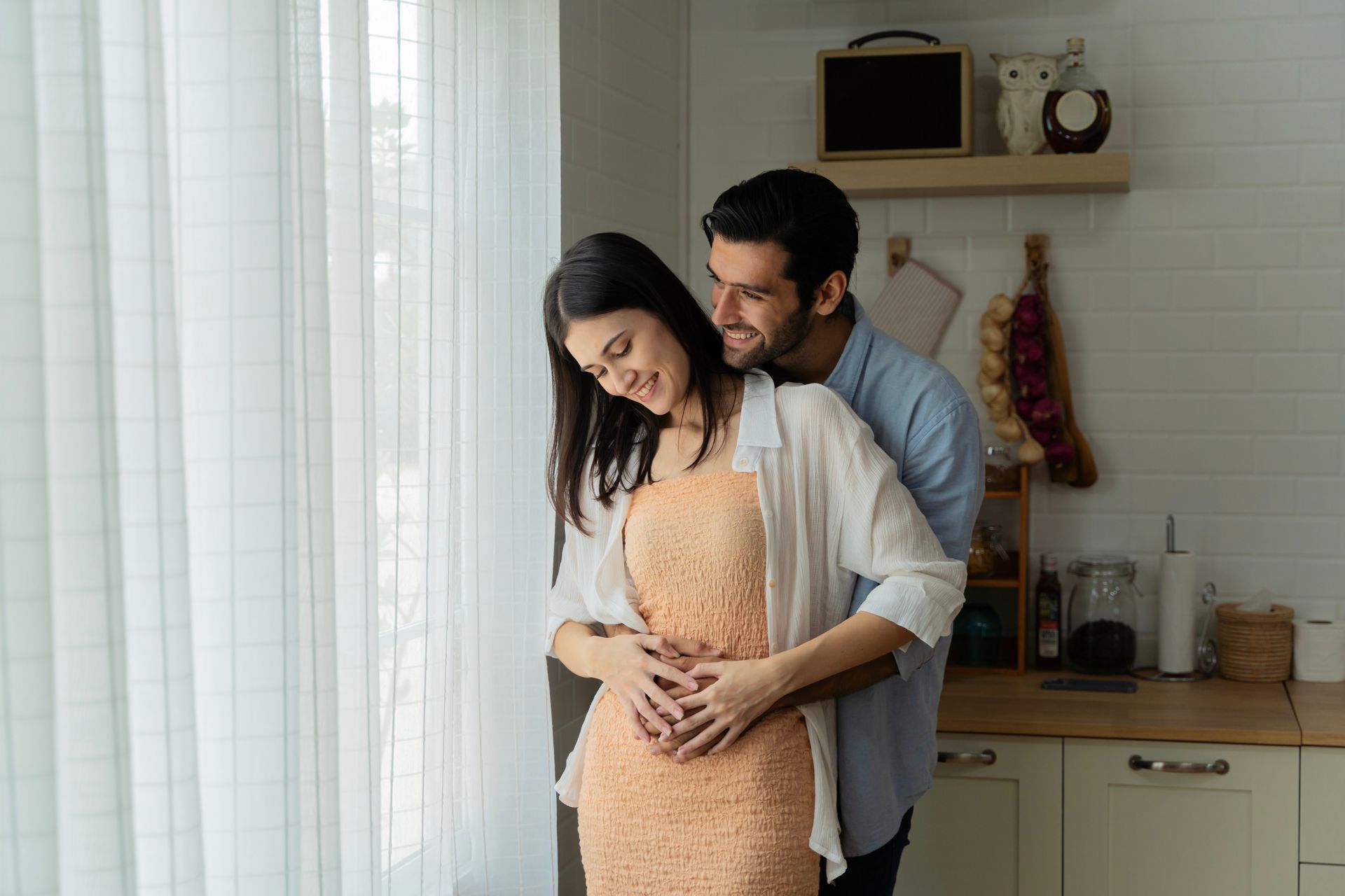 Un couple dans la cuisine, enlacé près de la fenêtre ; les bras de l'homme autour de la taille de la femme, souriant.