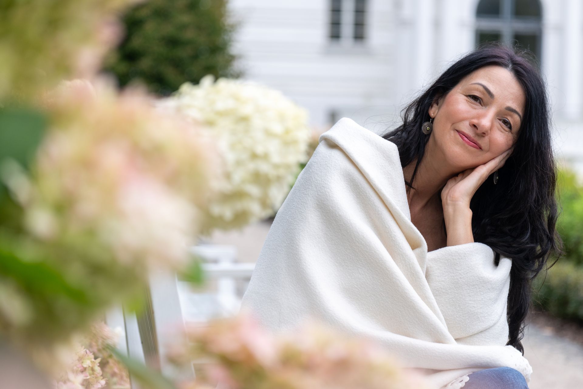 Une femme enveloppée dans une couverture blanche, souriante, est assise sur un banc devant un bâtiment blanc orné de grandes fleurs blanches.