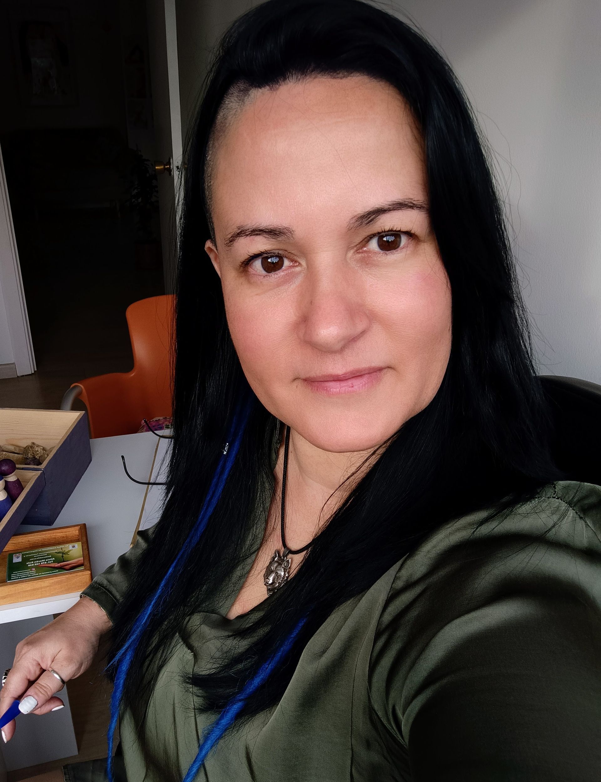 Mujer de cabello oscuro, con los laterales rapados, con camisa verde. Sonriente, en un espacio interior, con joyas visibles.