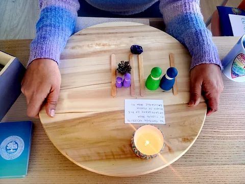 Persona sosteniendo una tabla de madera con muñecos de clavija, una vela y una nota sobre una mesa.