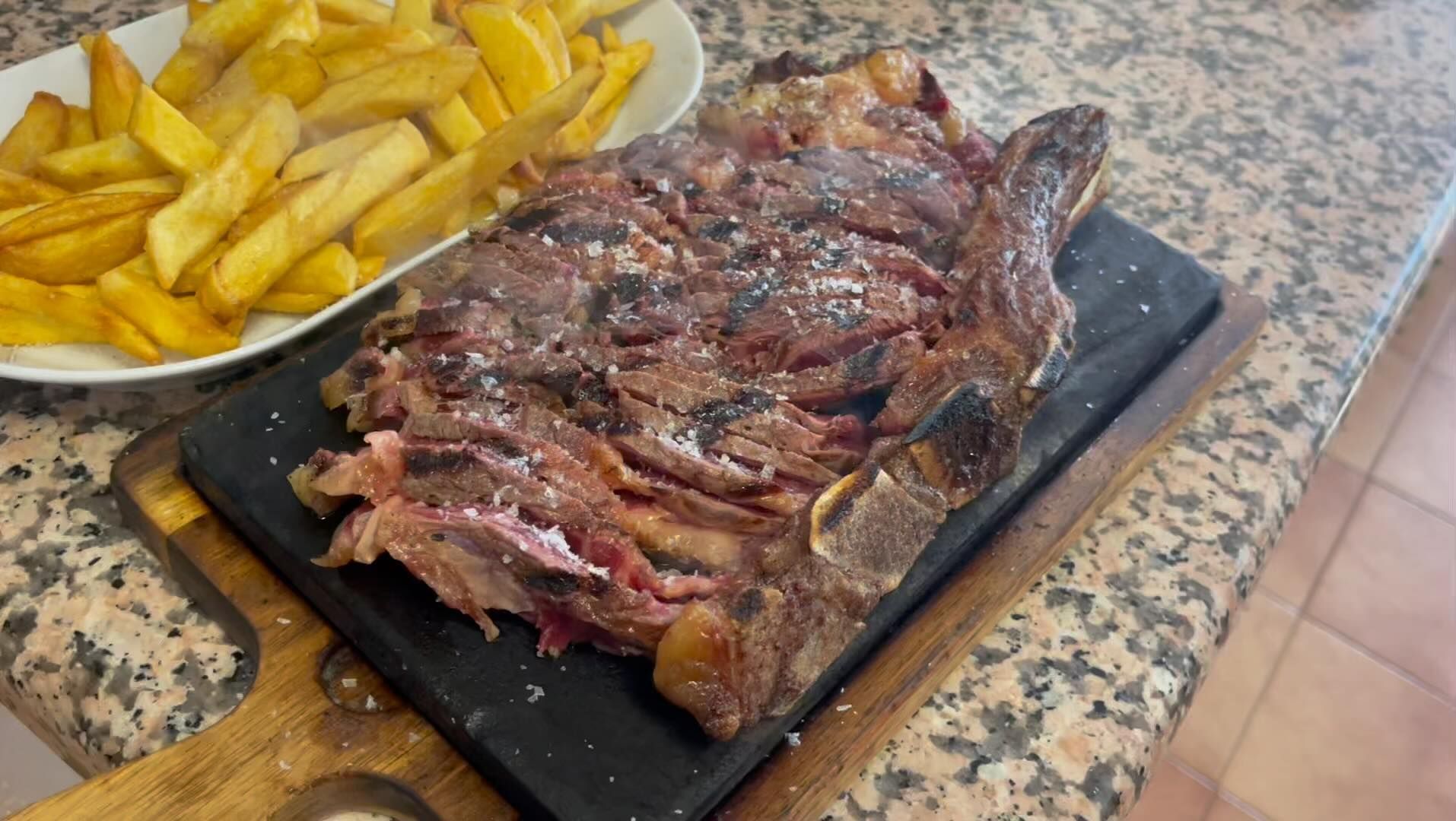 Un gran trozo de carne se encuentra en una tabla de cortar al lado de un plato de papas fritas.