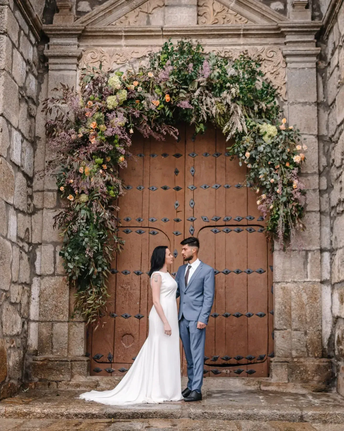 Una novia y un novio están de pie frente a una puerta de madera decorada con flores.