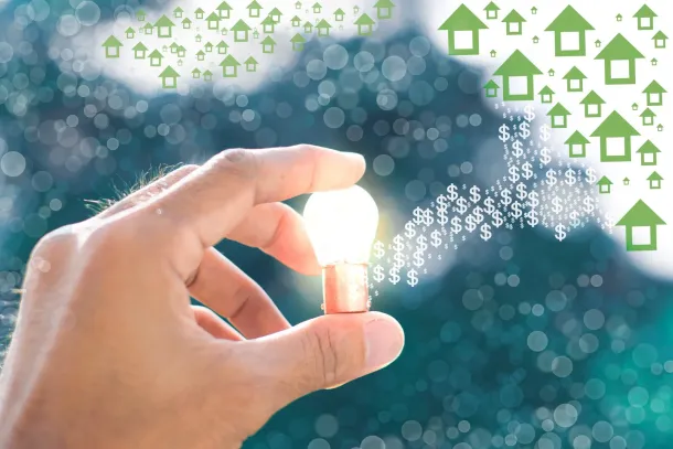 Una mano sostiene una bombilla brillante con iconos verdes de casas flotando al fondo.
