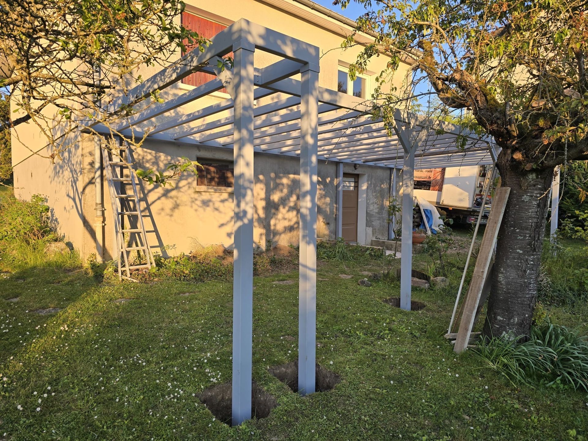Structure de pergola en bois partiellement construite dans une cour herbeuse à côté d'une maison.