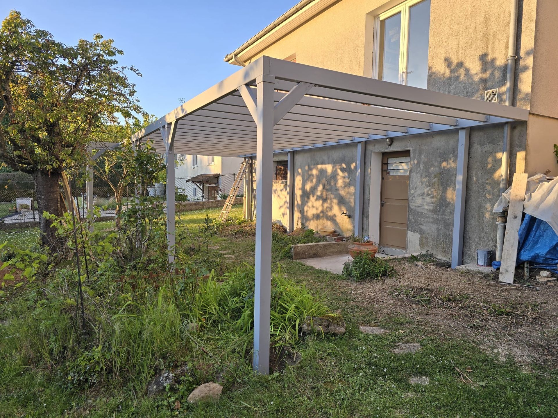 Terrasse arrière avec une pergola grise à côté d'une maison beige, entourée de pelouse et de plantes de jardin.
