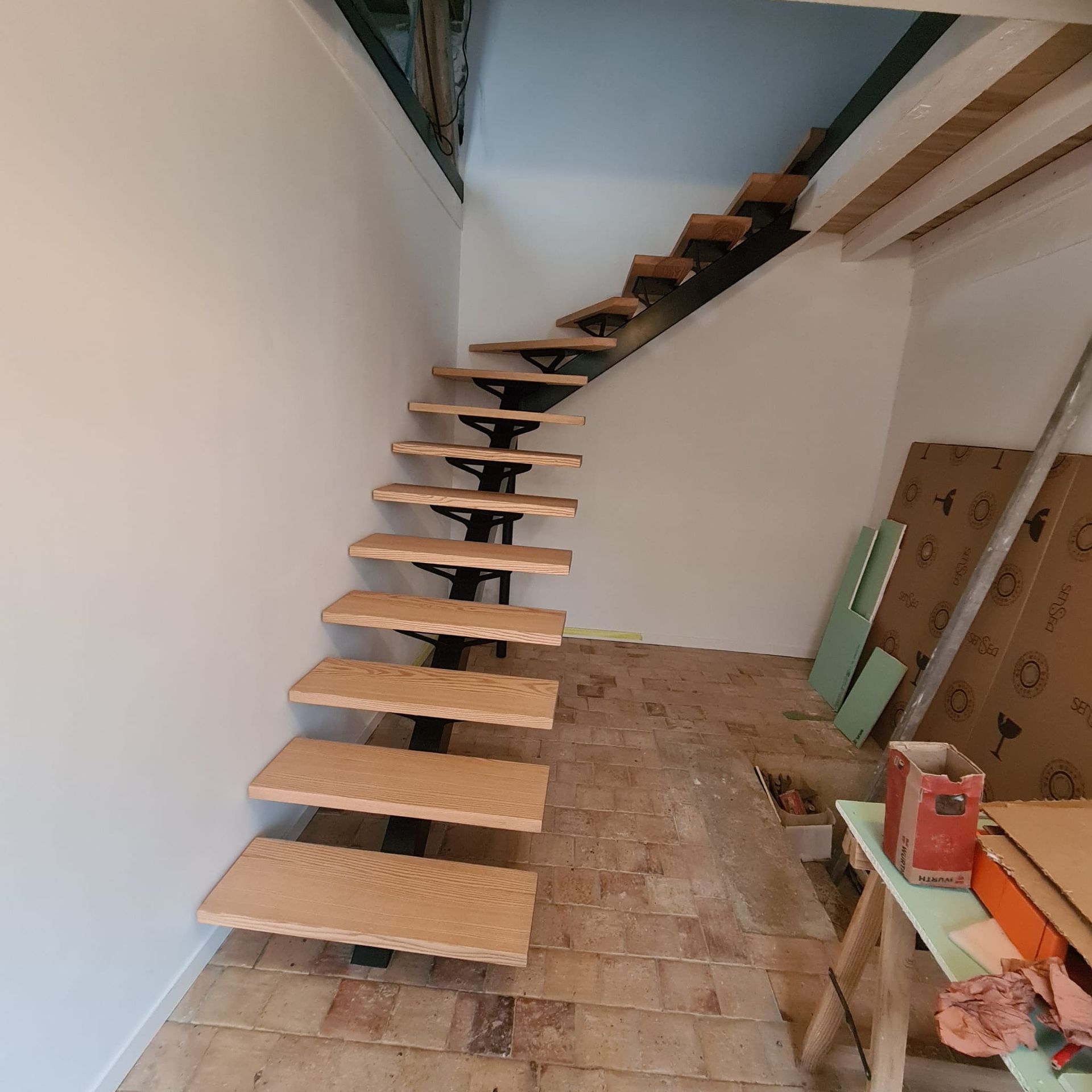 Escalier flottant en bois courbé dans une pièce inachevée aux murs nus et aux outils éparpillés au sol.