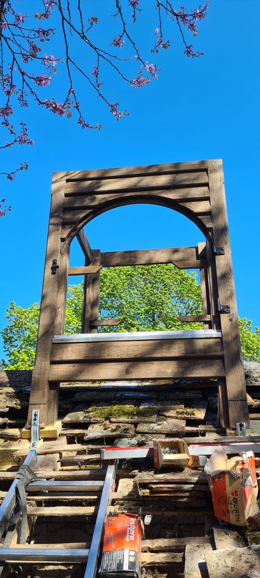 Une structure en bois patinée par le temps, sous un ciel d'un bleu éclatant, entourée de bourgeons rouges et de verdure.