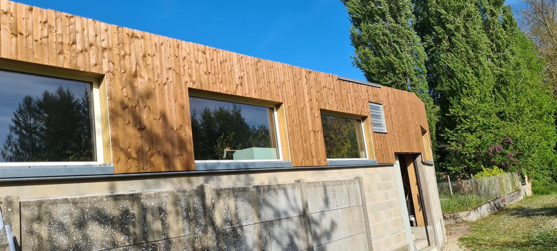 Bâtiment moderne revêtu de bois avec de grandes fenêtres, jouxtant un mur de pierre et des arbres sous un ciel bleu.