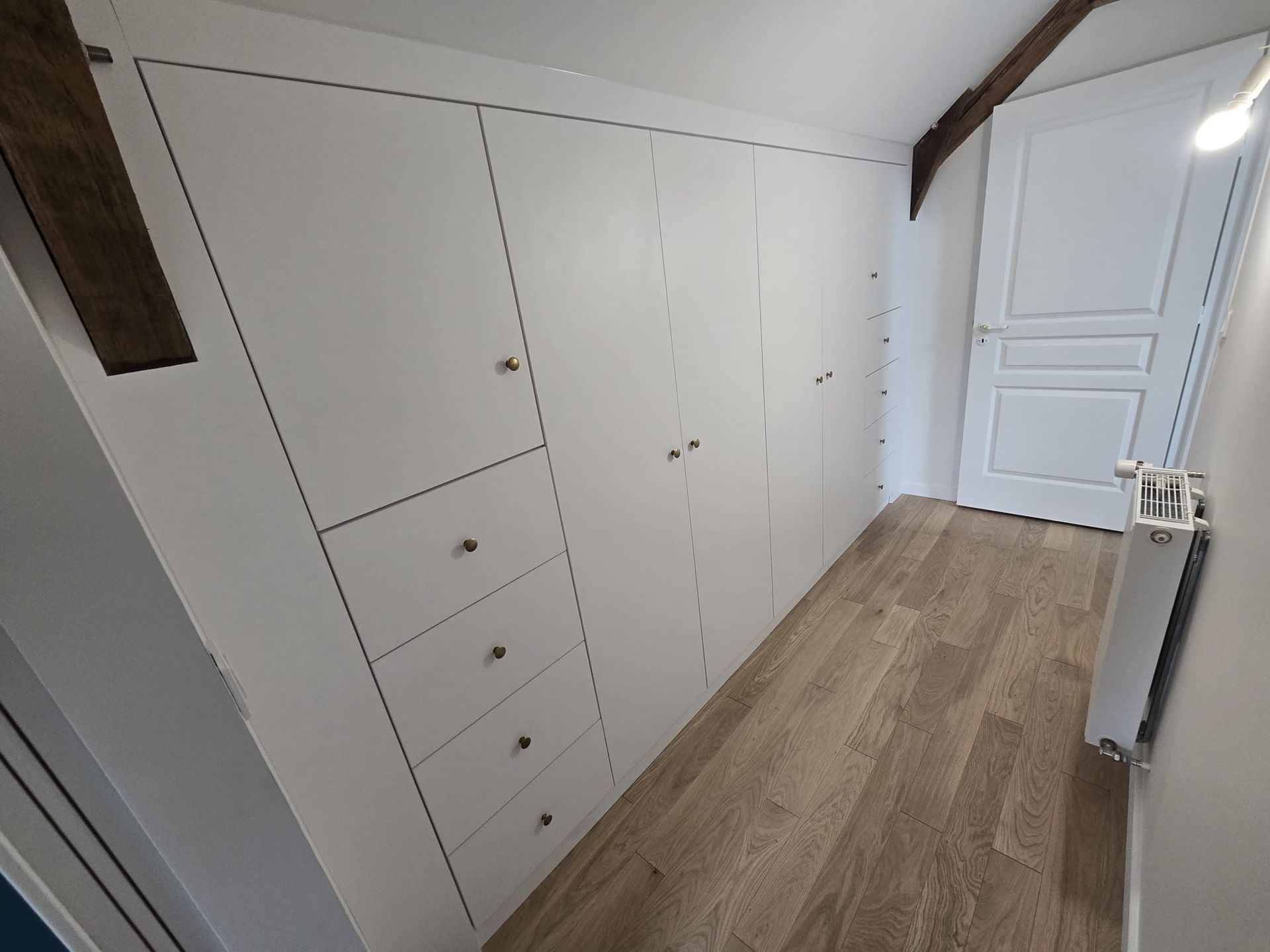 Placard mural blanc intégré dans une pièce mansardée avec parquet et radiateur près de la fenêtre.