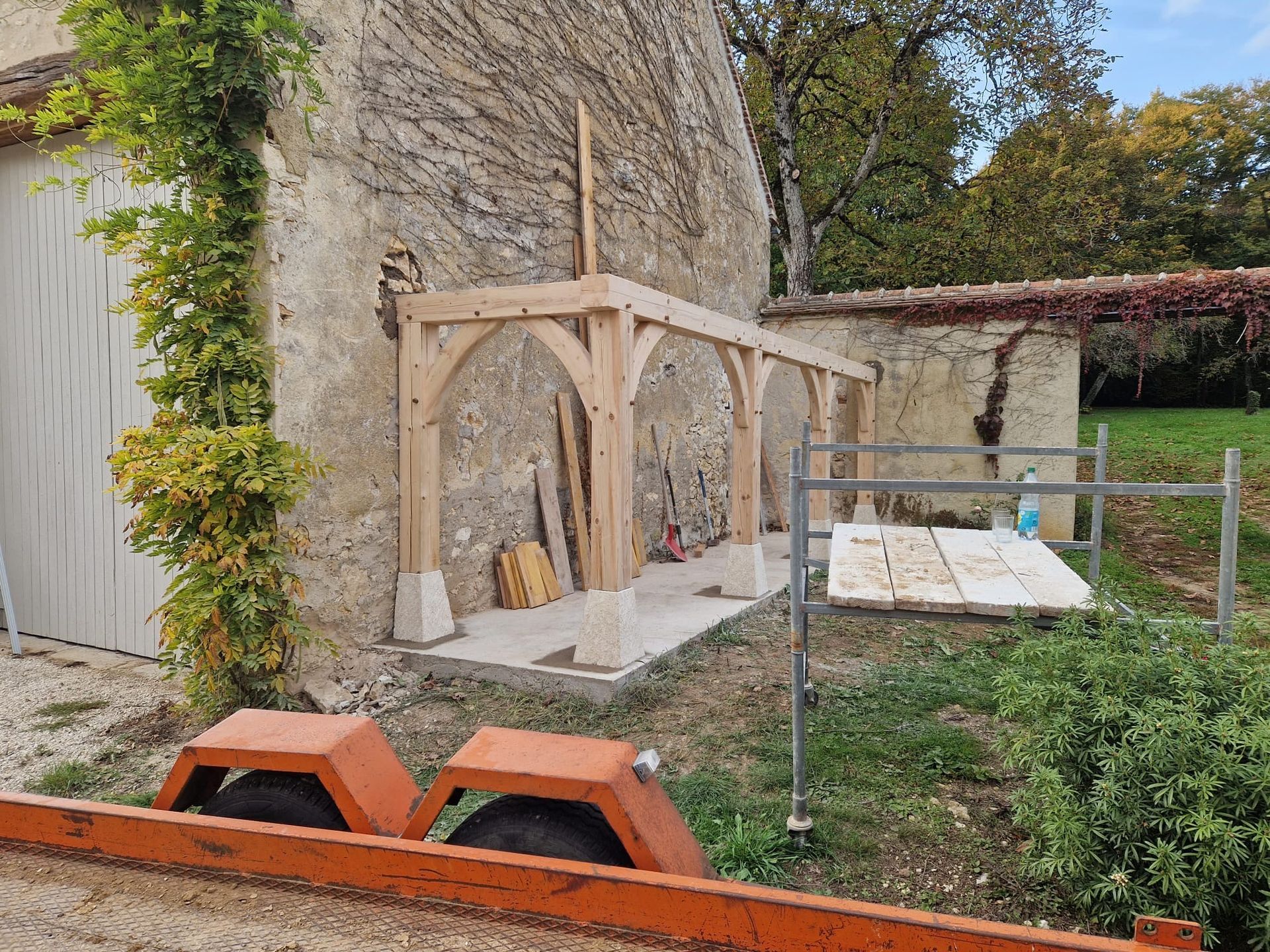 Des abris en bois sont adossés à un muret en pierre dans une cour herbeuse, avec des outils.