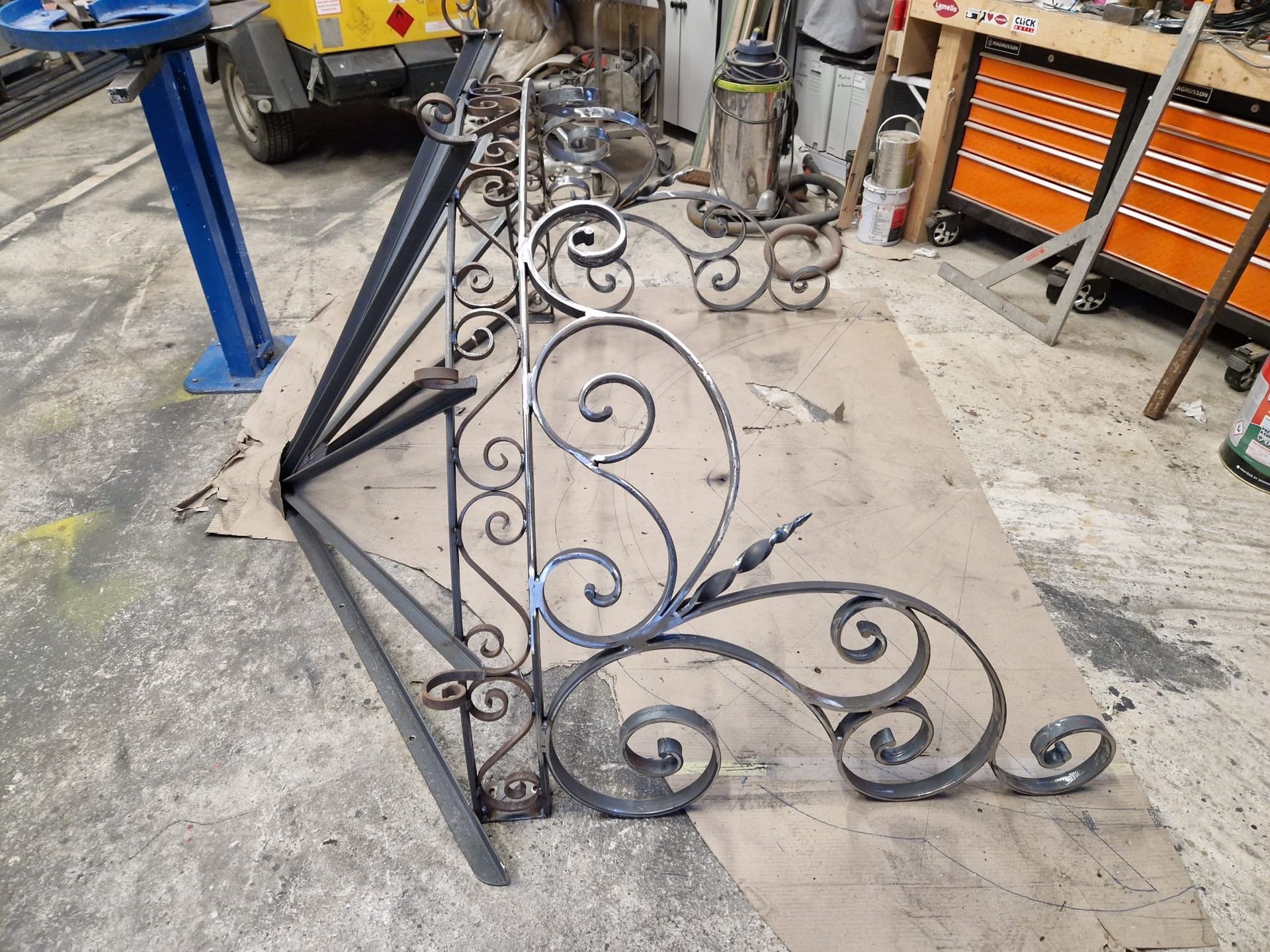 Des sections de rampe d'escalier en fer forgé noir ouvragé, posées sur le sol d'un atelier à côté d'outils et d'équipements.