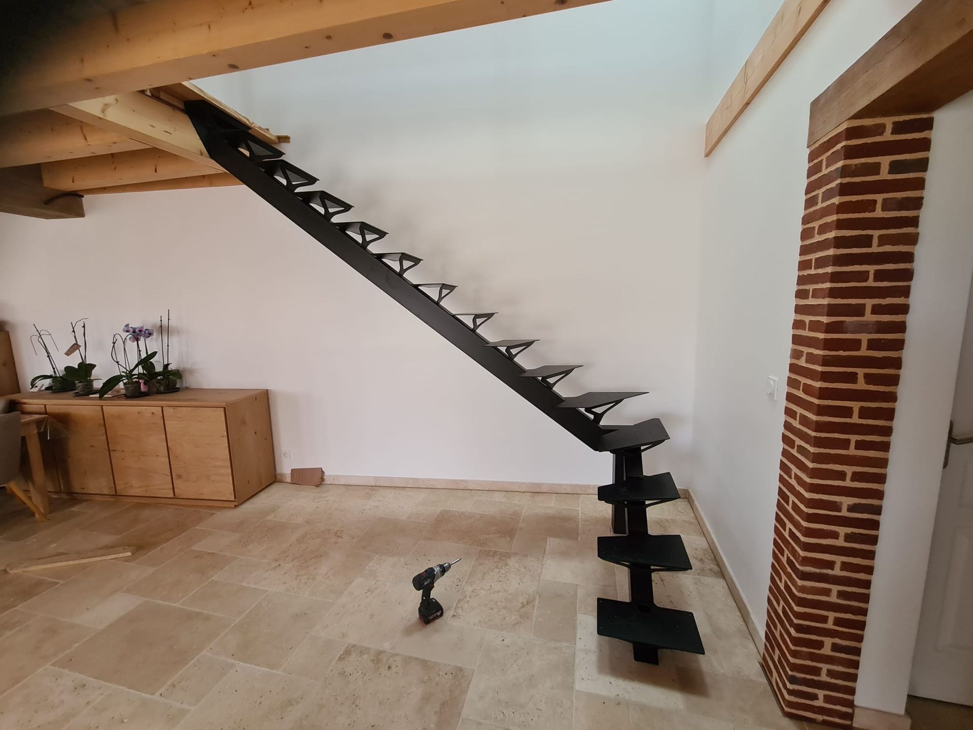 Intérieur moderne avec un escalier flottant noir, une colonne en briques et des poutres en bois au-dessus d'un sol carrelé.